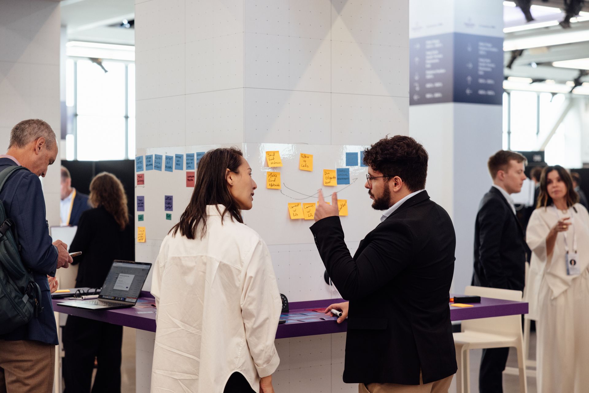 Two people converse at a display with sticky notes, others in background.