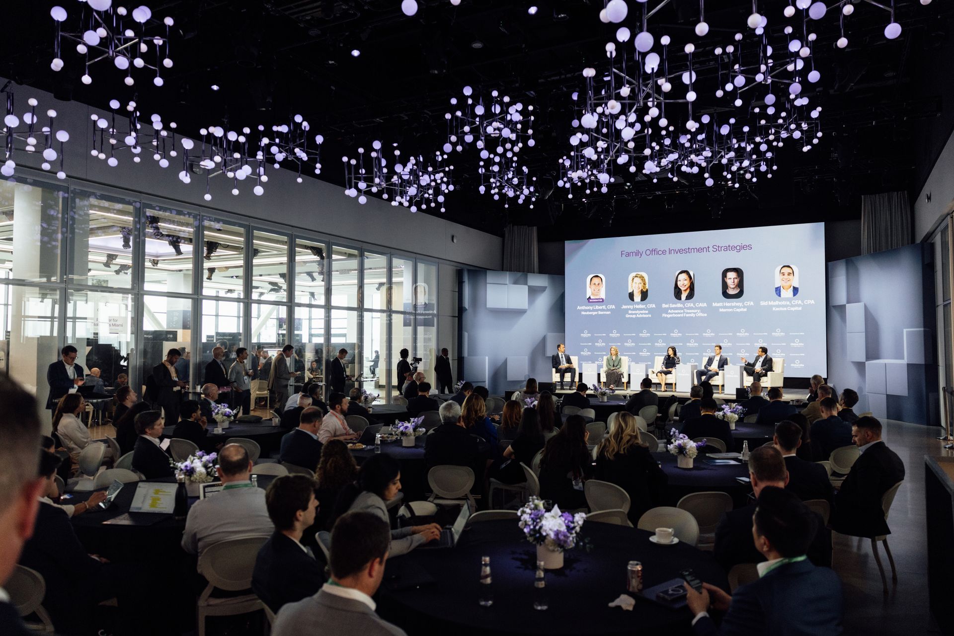Conference hall with panel discussion, audience seated at tables, modern architecture, and overhead lighting.