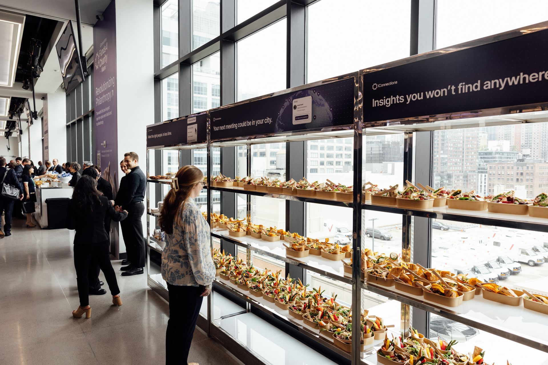 People at an event, viewing food displayed on shelves, with a city view through large windows.