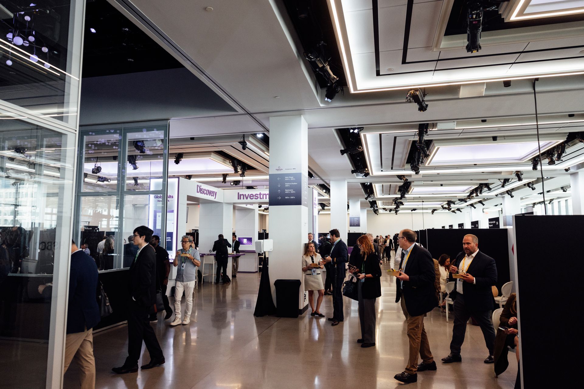 People in business attire at a conference, hall with bright lights, glass walls, and glossy floor.