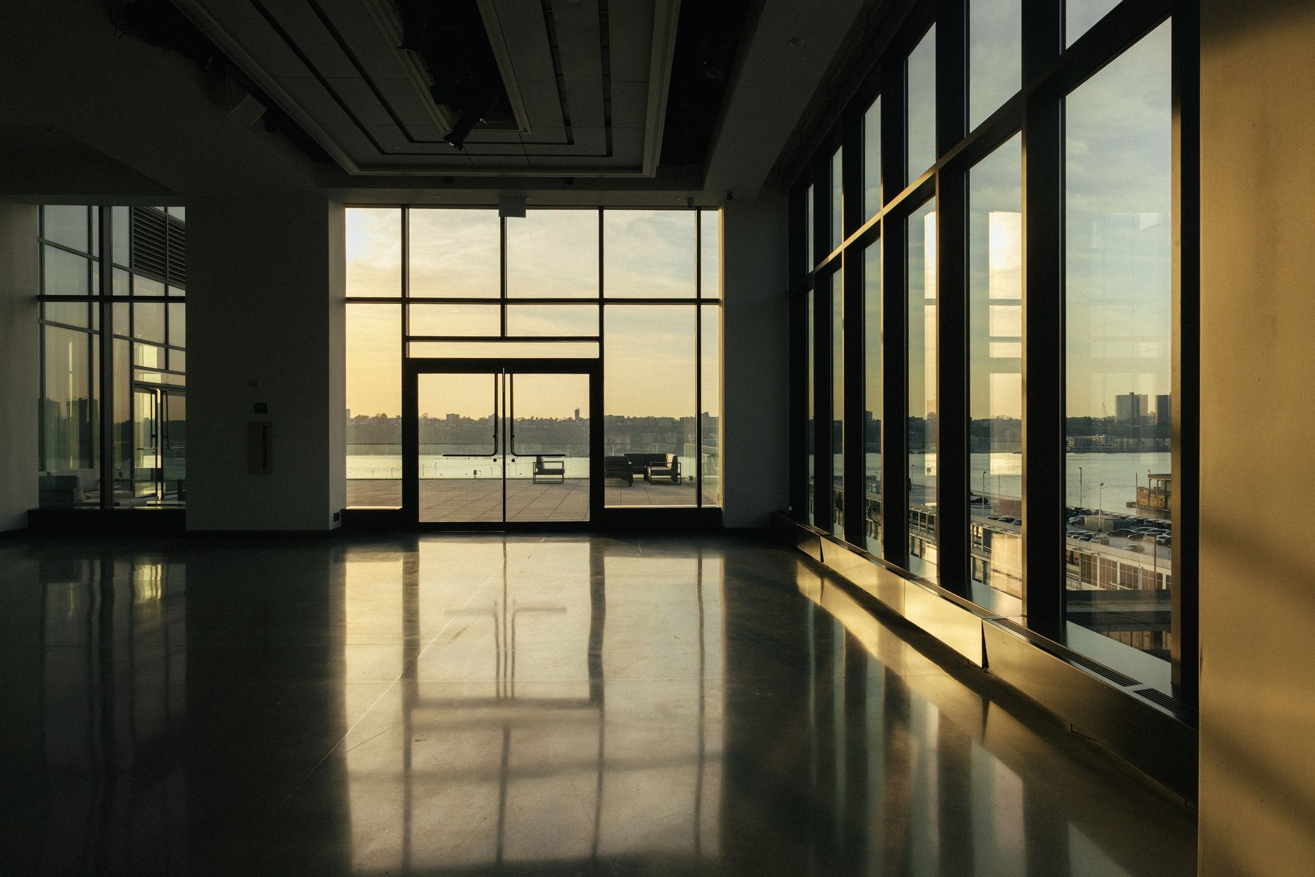 Empty, sunlit room with large windows overlooking a waterfront at dusk.
