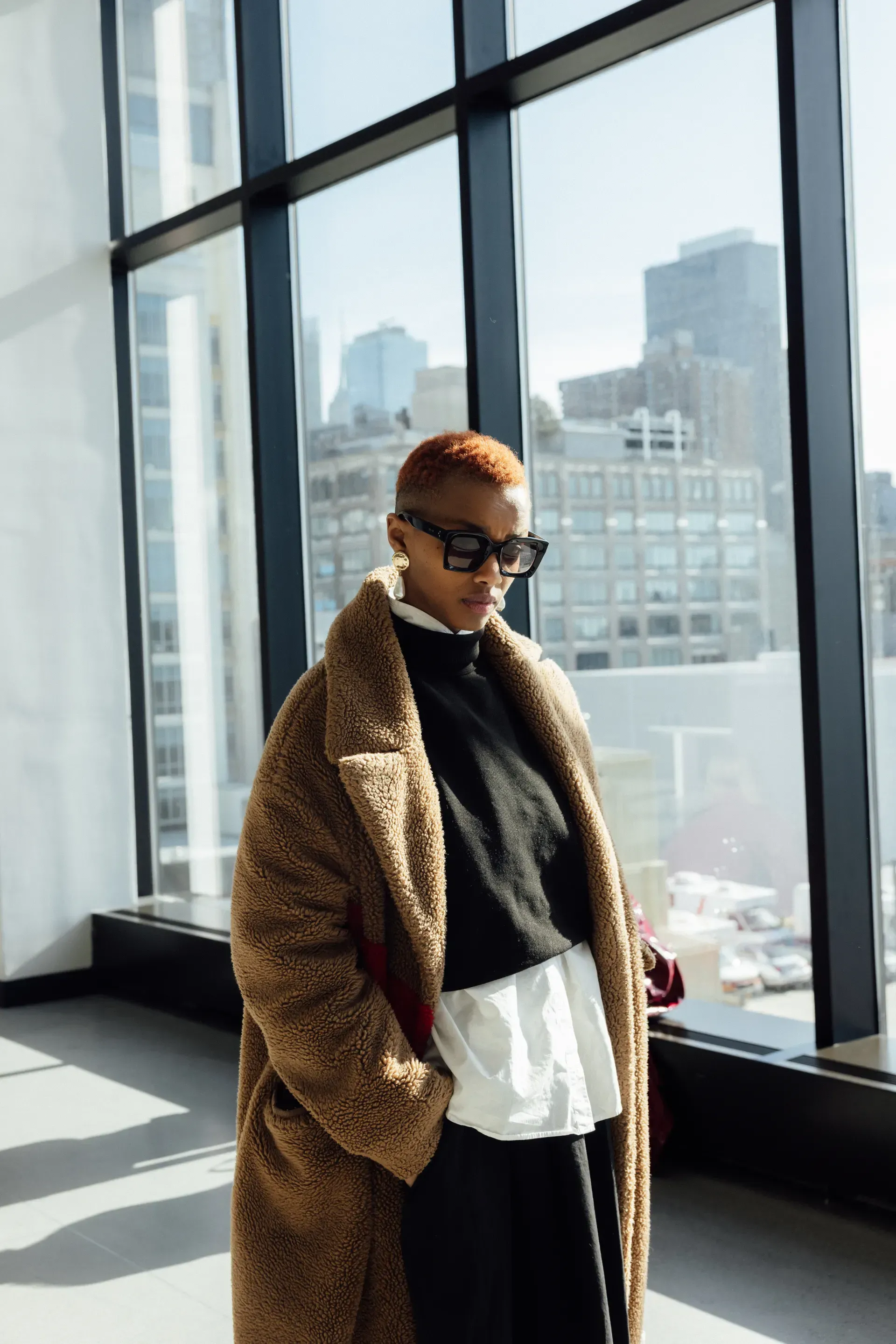 A woman wearing sunglasses and a teddy bear coat is standing in front of a window.
