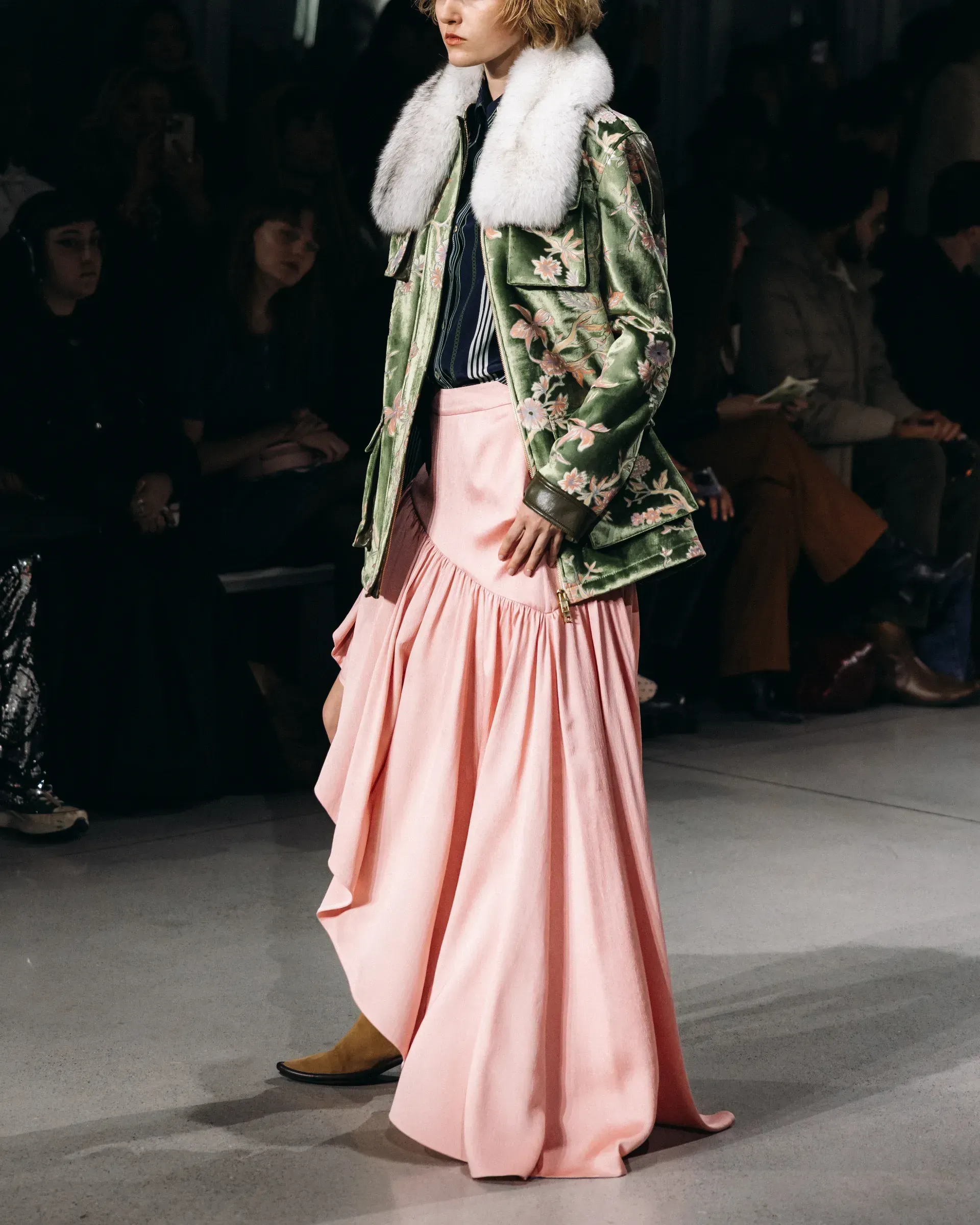 A woman is walking down a runway wearing a pink skirt and a green jacket