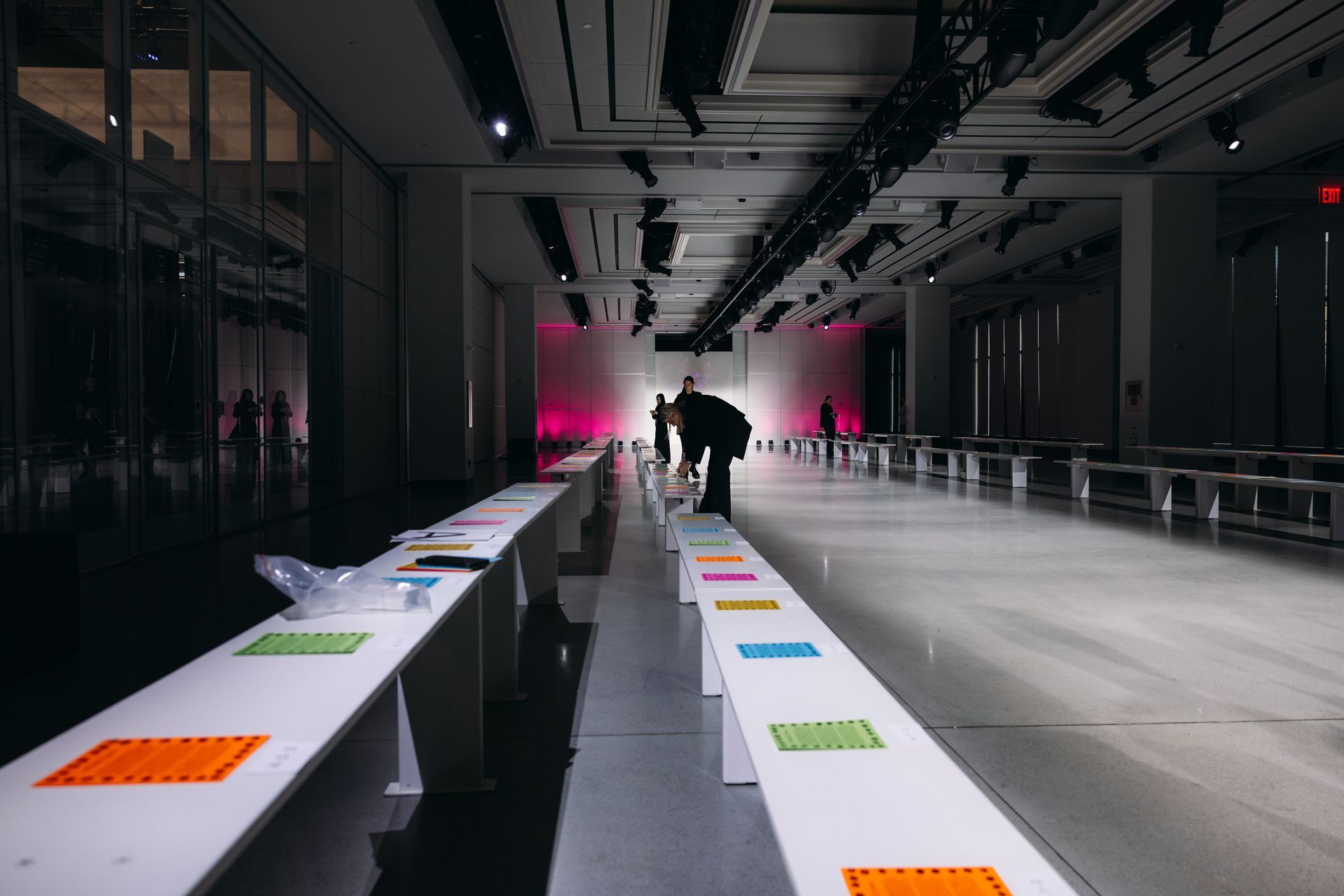 Empty fashion runway with rows of white benches, lit by overhead lights; a few people are in the background.