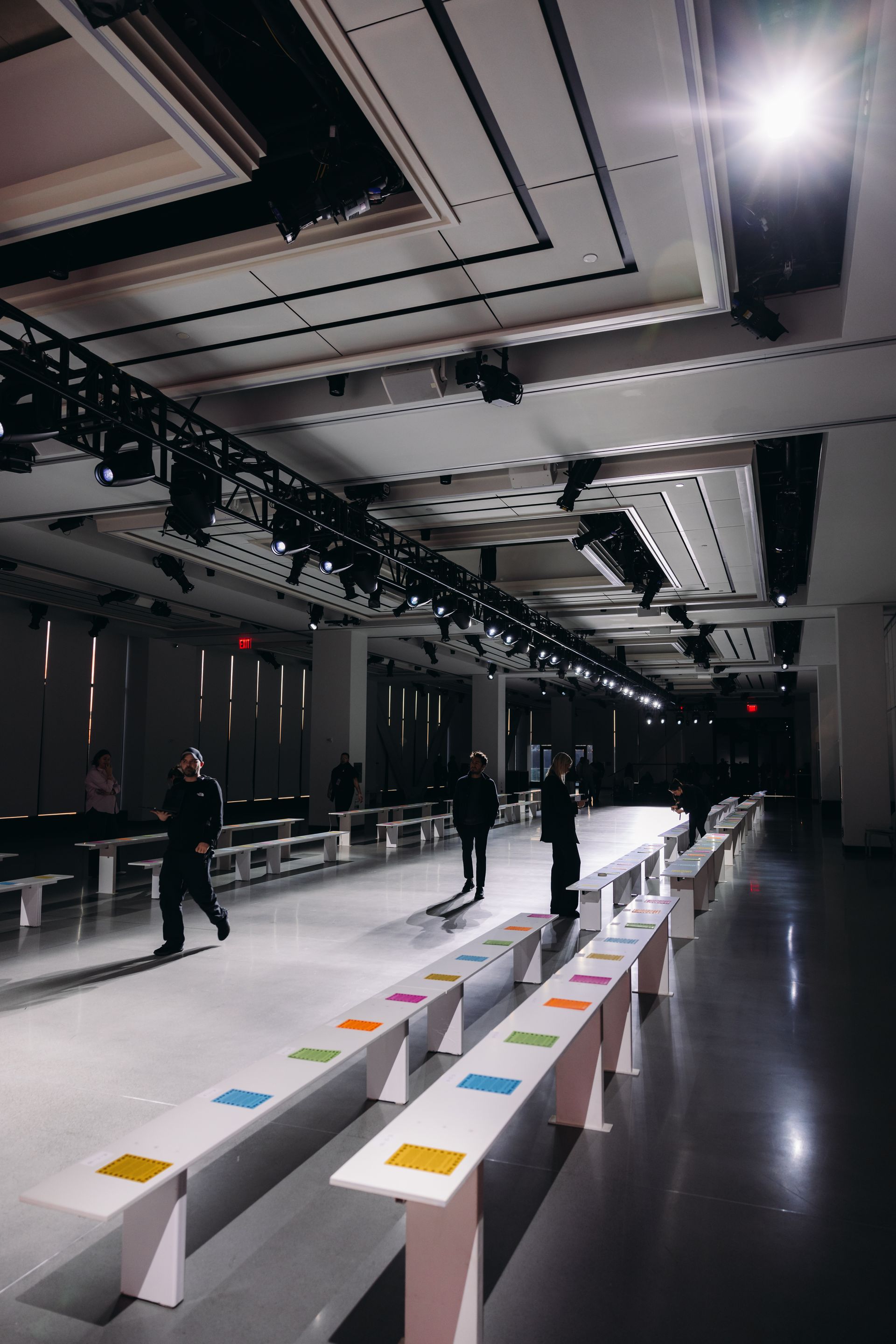 Empty fashion show venue with rows of seats, overhead lighting, and people walking.