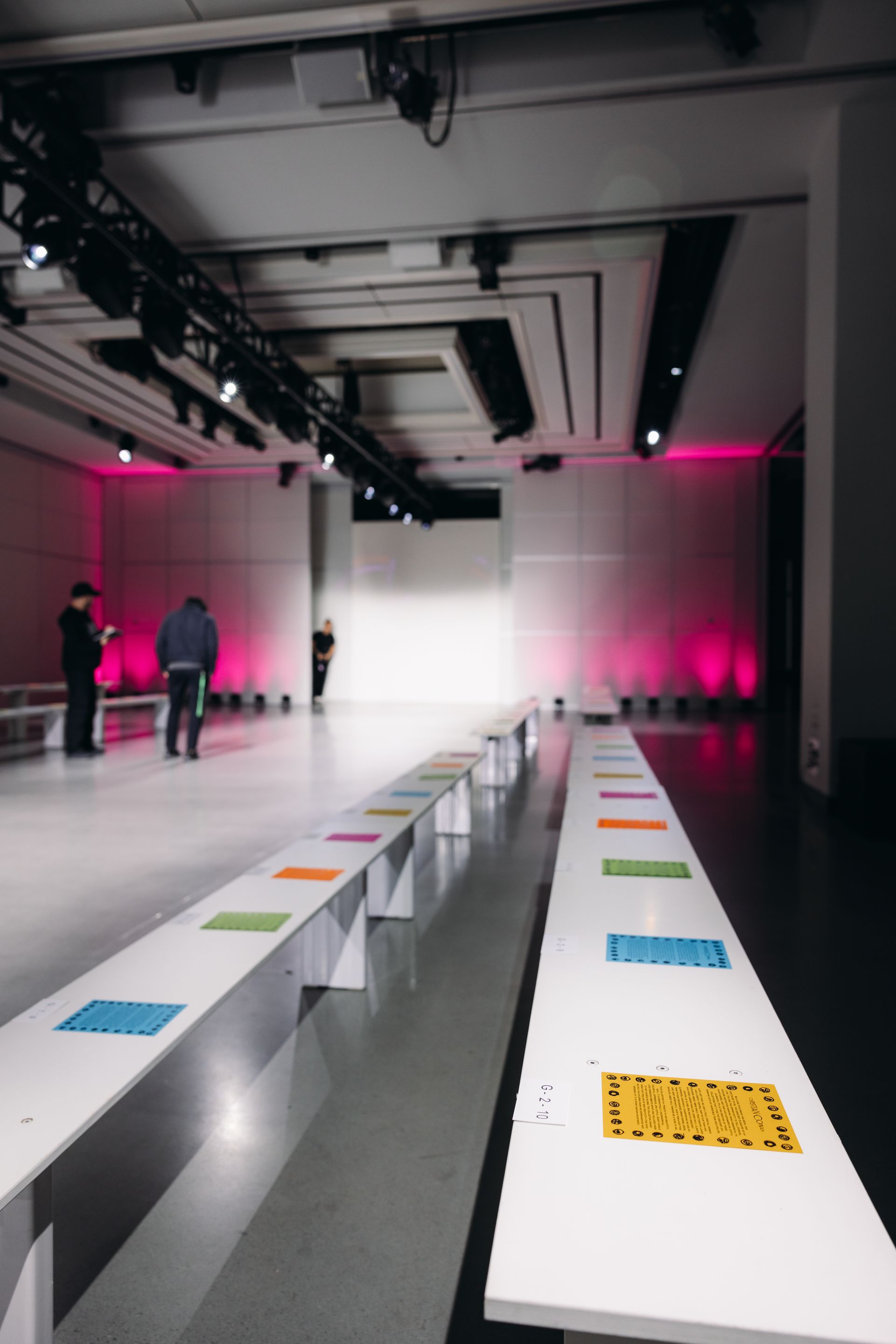 Fashion show venue with rows of white benches with colorful seat assignment cards, runway, pink lighting.