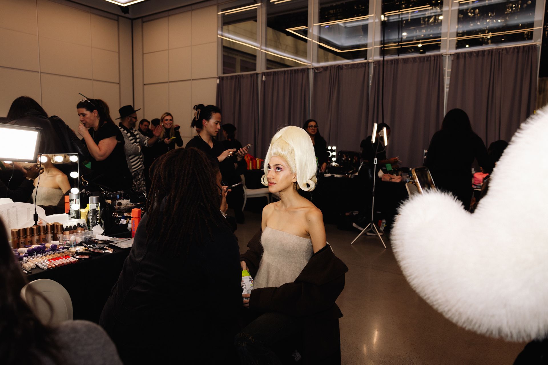 A model with a tall wig in a makeup room, surrounded by makeup artists and equipment.