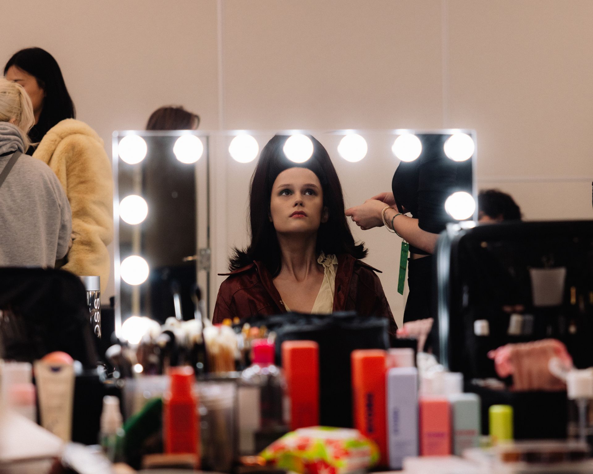 Woman in costume looks in mirror, getting hair styled in a backstage setting with makeup and lights.