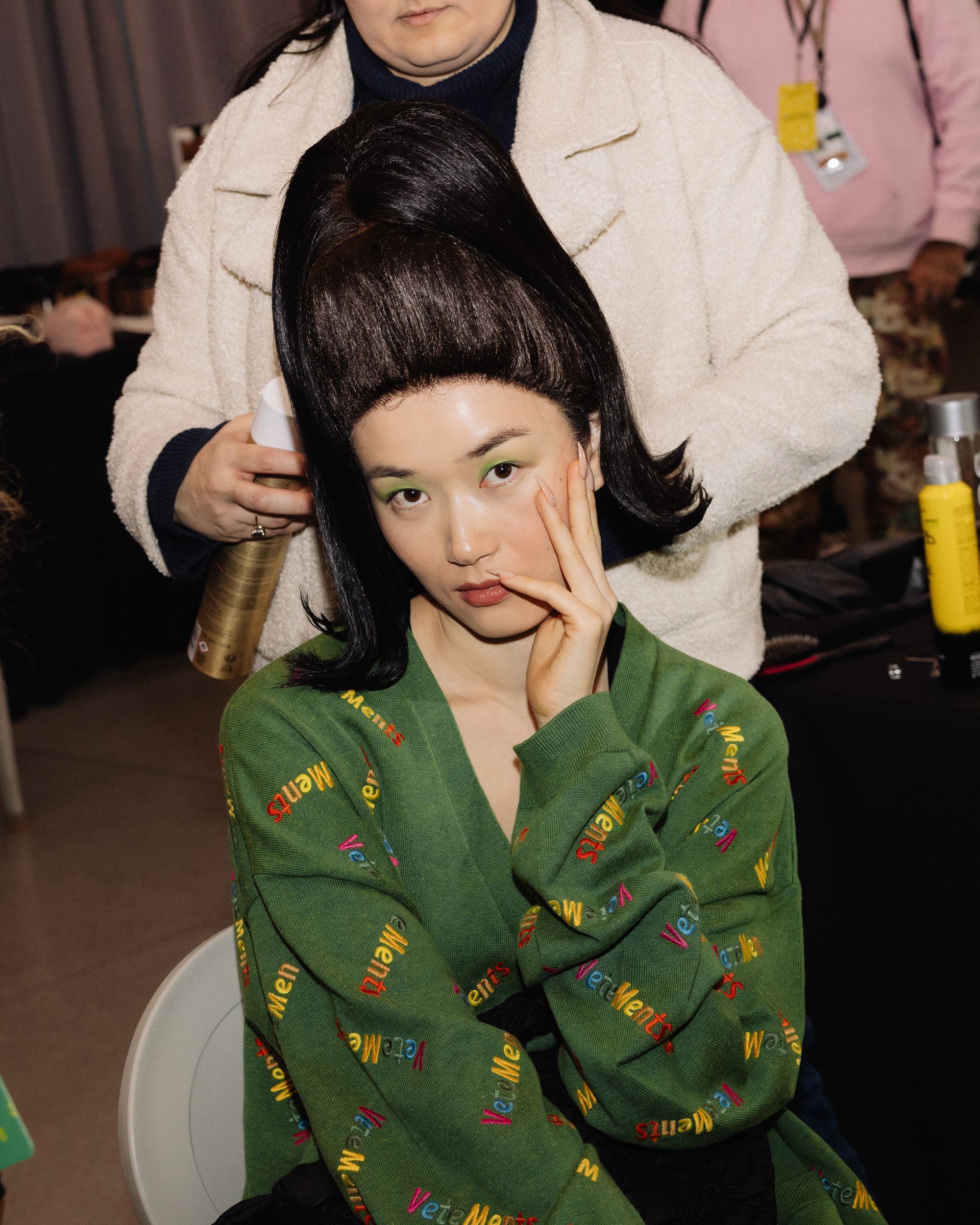 Woman in a green robe with large black wig, being styled. A person in a white jacket is behind her.