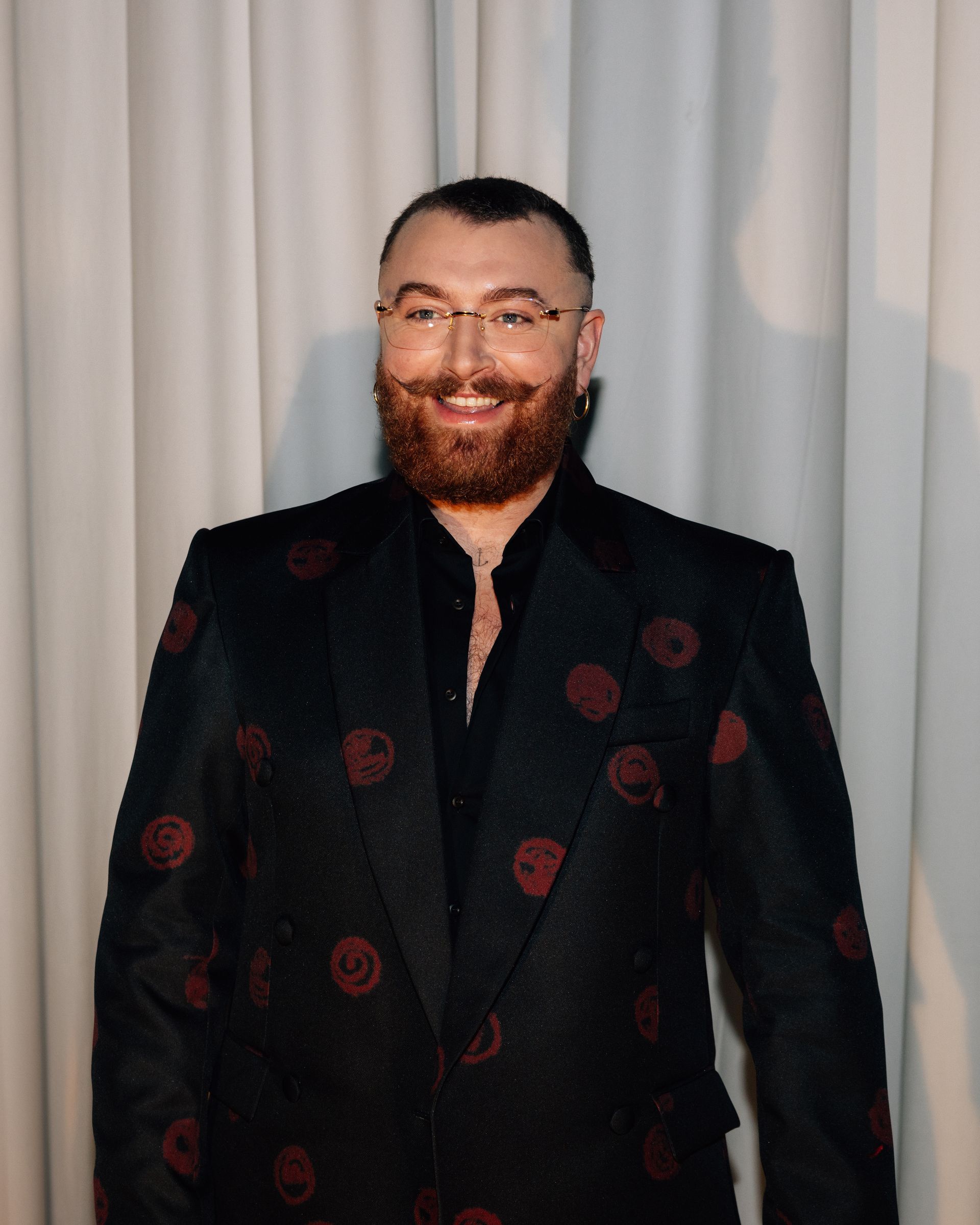 Sam Smith smiles in a black suit with red embellishments, glasses, and a full beard.