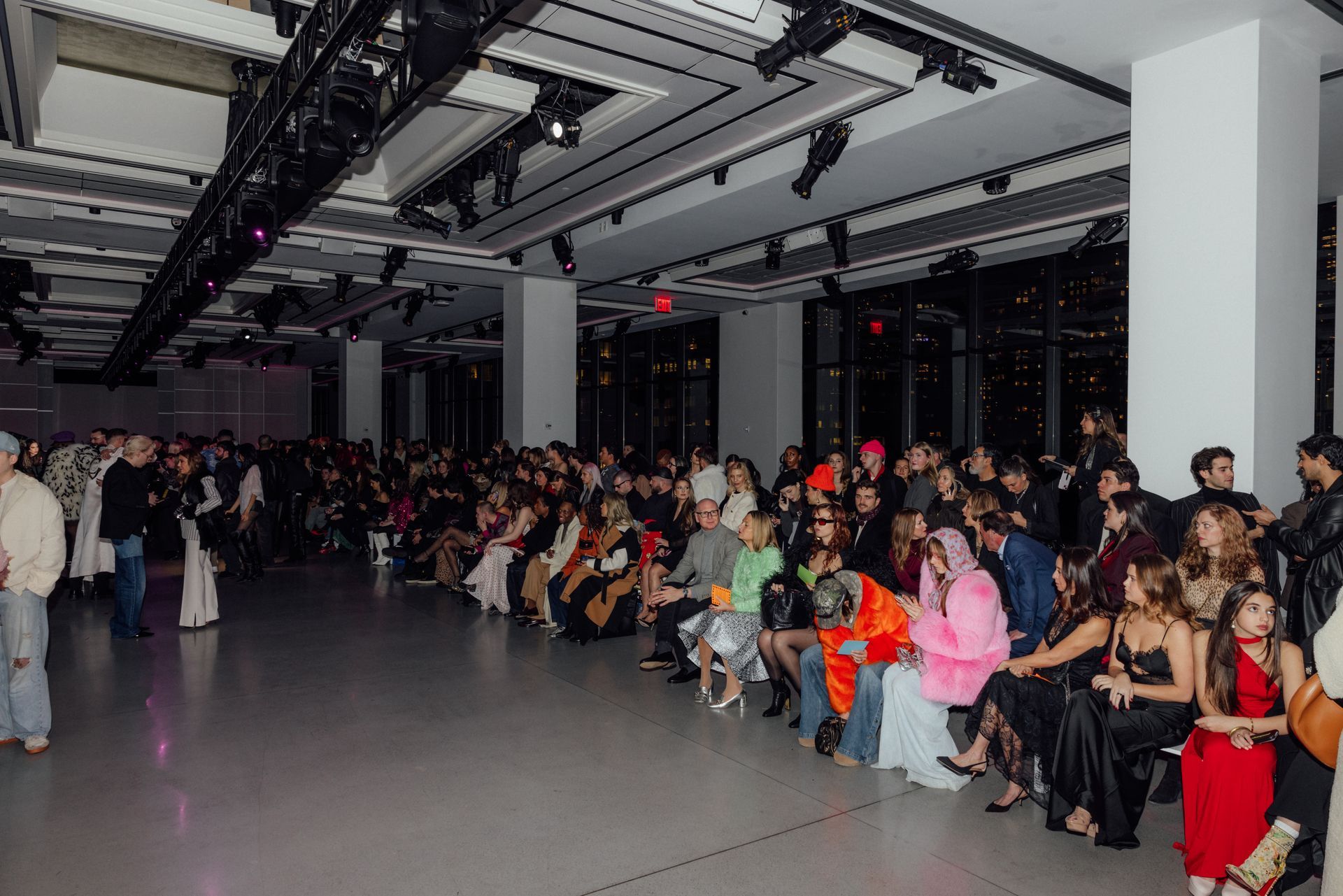 Fashion show audience in an interior space. People seated, watching a runway; nighttime view through windows.