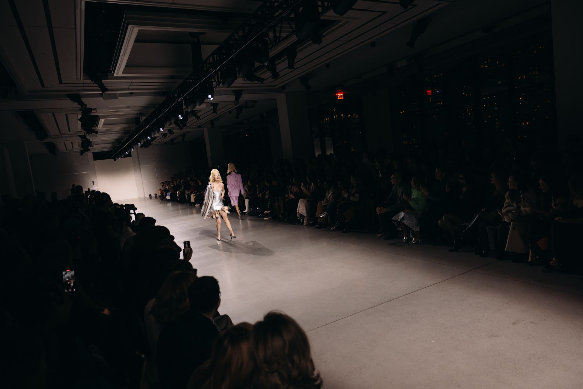 Fashion show runway with model under bright lights, audience on sides.