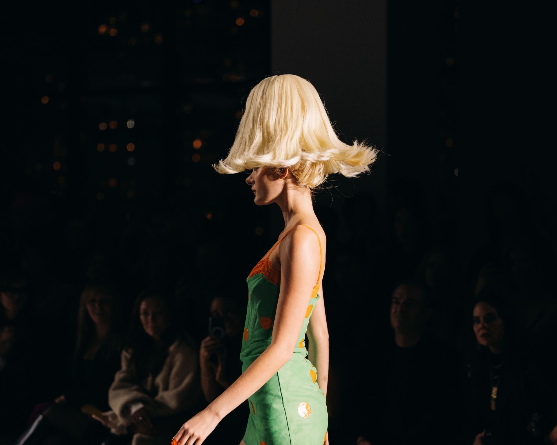 Model in a green dress with a unique mushroom-shaped blonde wig walks a runway in a darkened space.