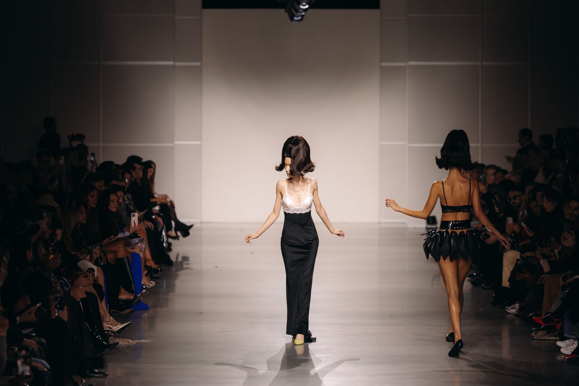 Models walking the runway at a fashion show, audience seated in the background.