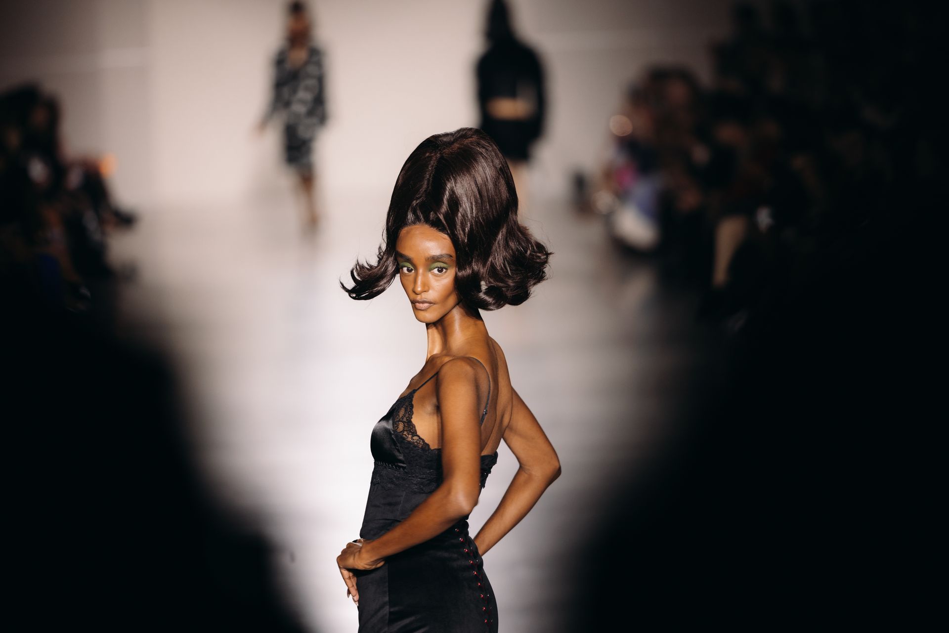Model on runway wearing black lingerie and large, dark updo; fashion show.
