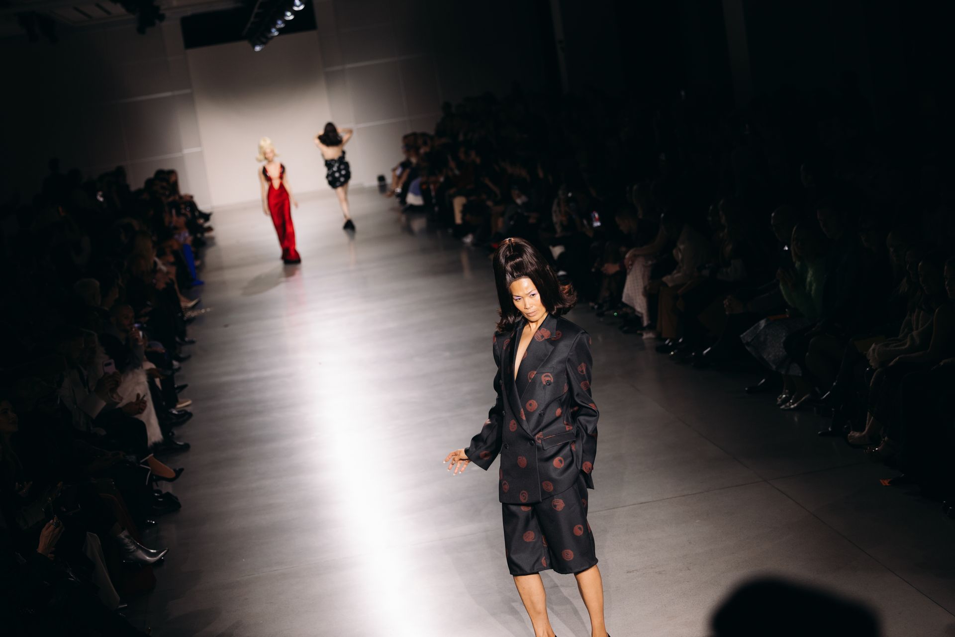 Fashion show runway with models in dark clothing; audience on the sides.