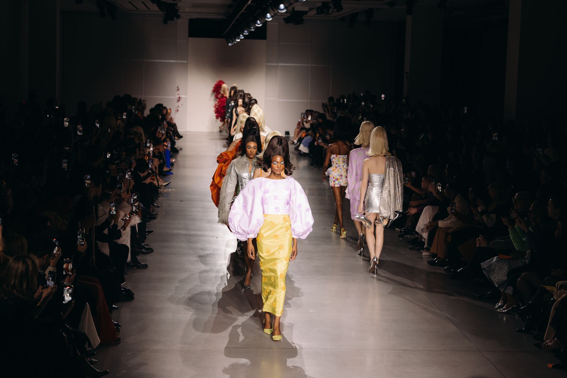 Fashion models walk down a runway, showcasing colorful outfits. Audience watches from the sides, illuminated by stage lighting.