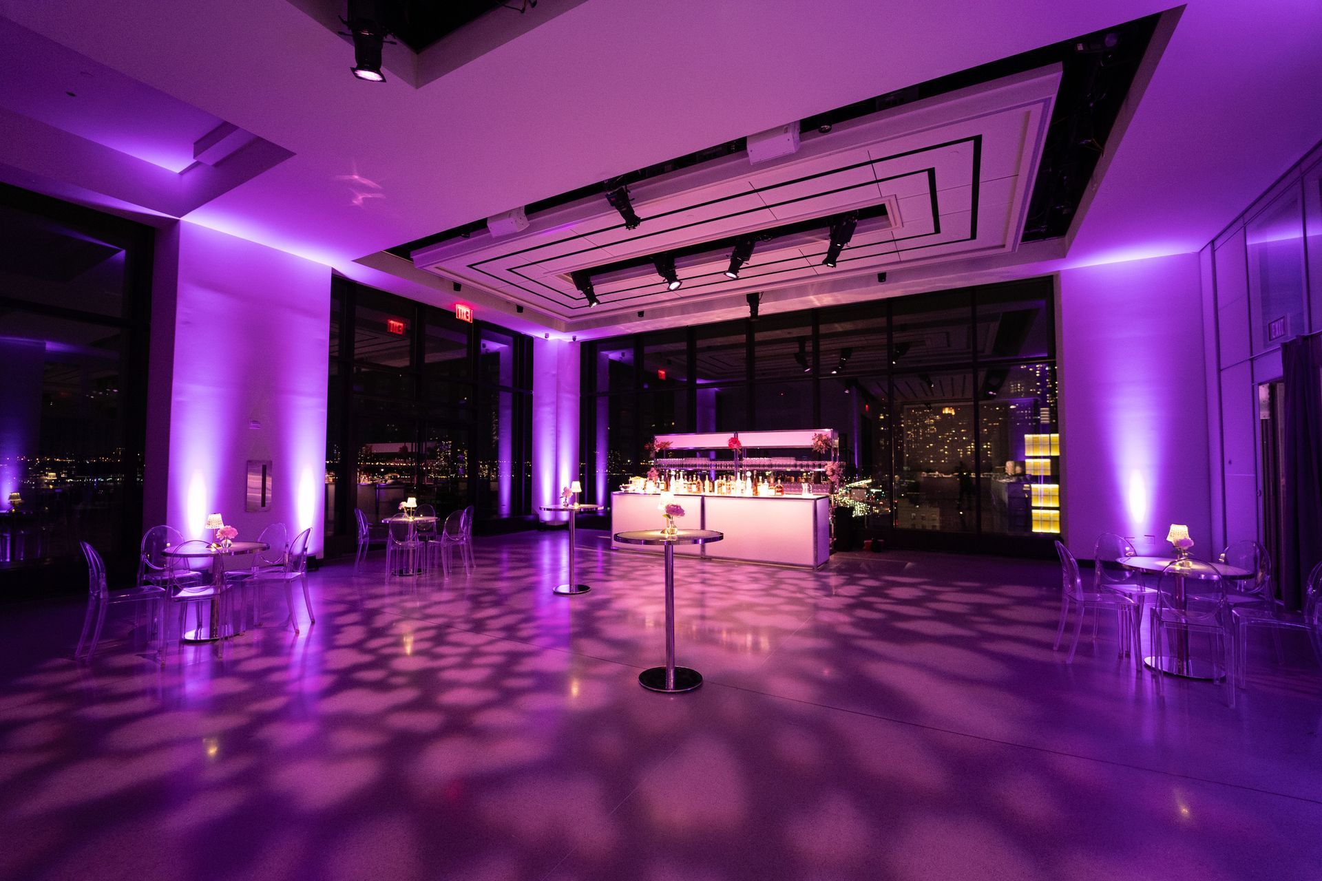 A modern event space lit with purple lights, tables and bar set up, large windows.