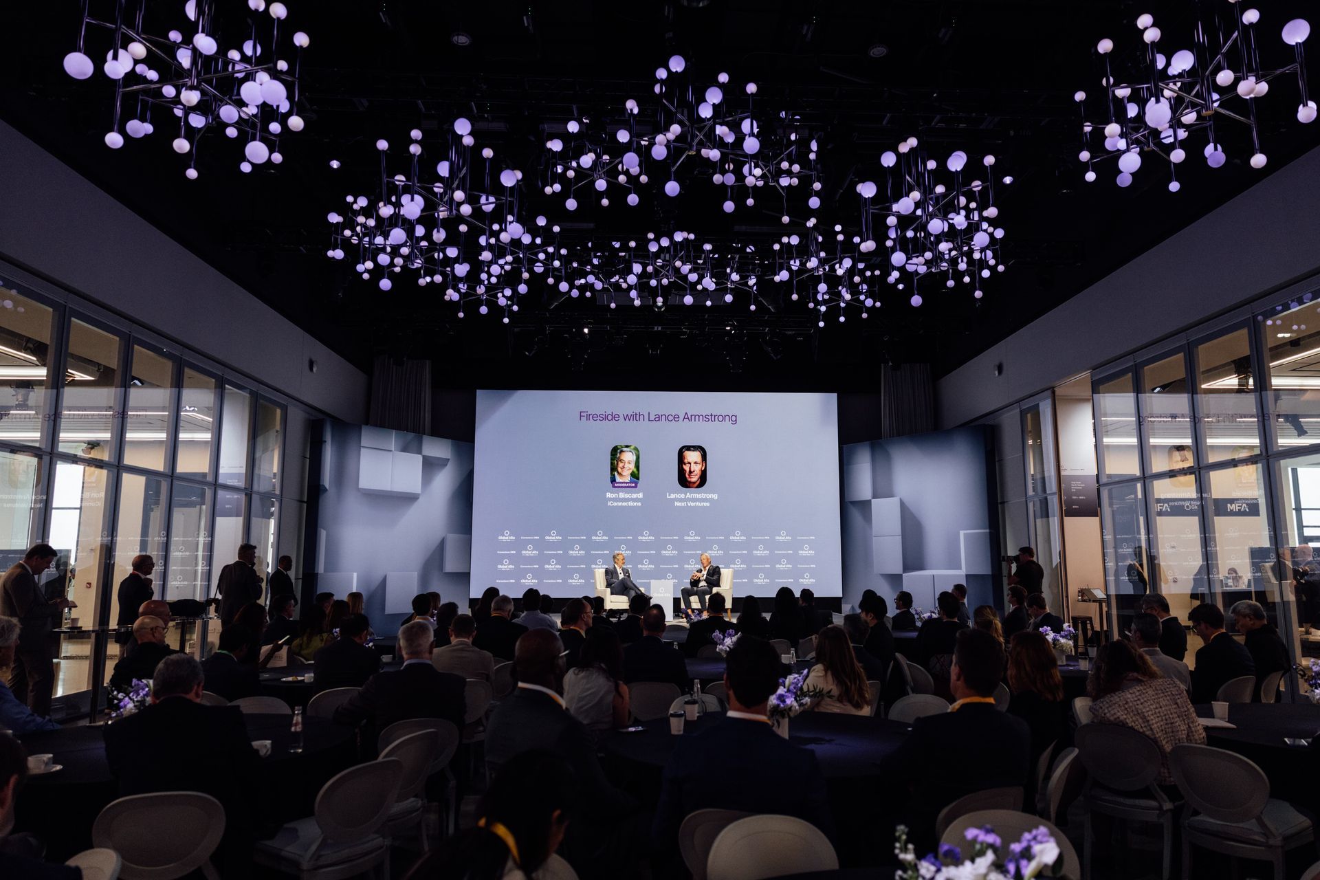Conference with speakers on stage, audience seated. Purple lights hang from the ceiling.