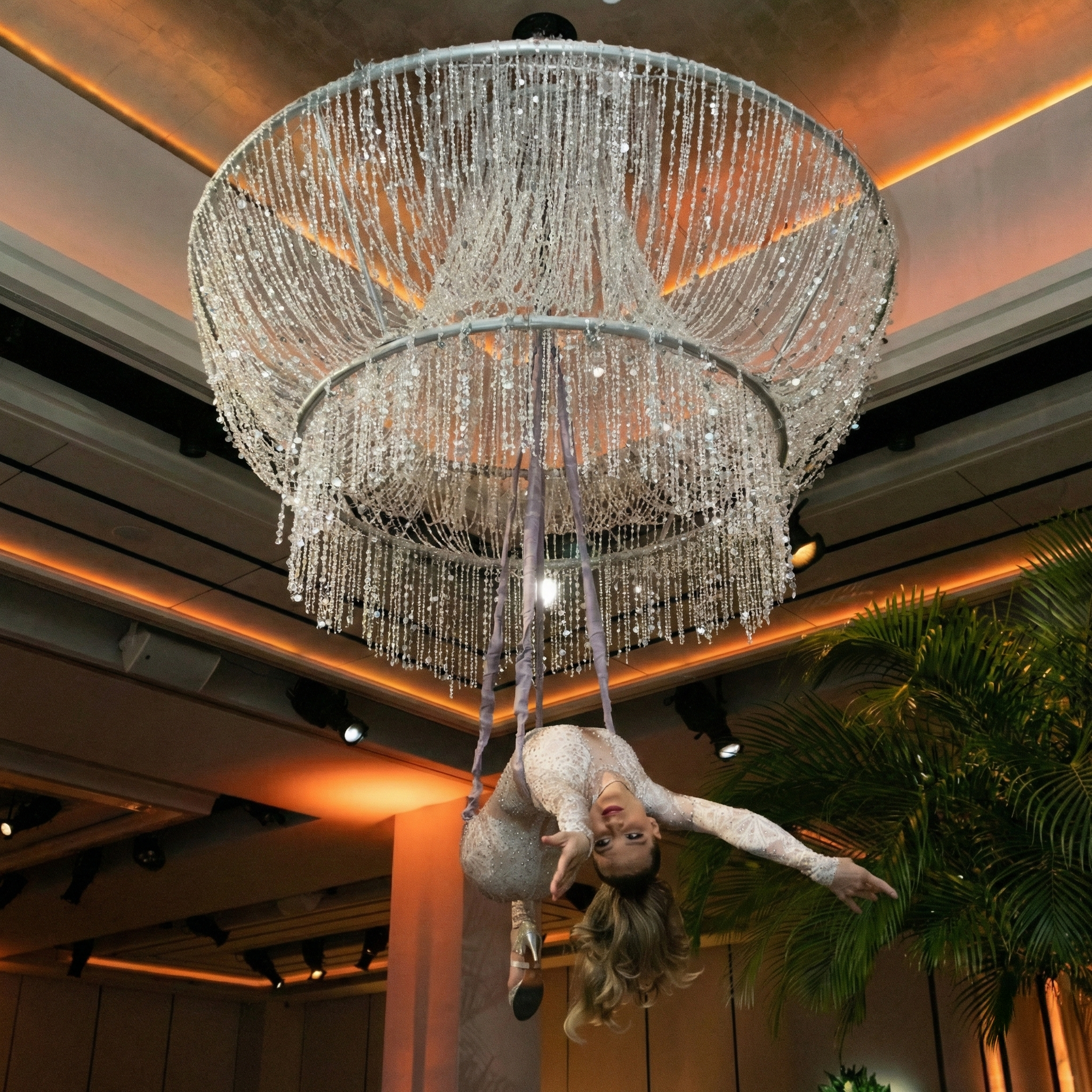 Woman in aerial silks suspended below a large crystal chandelier, arms outstretched.