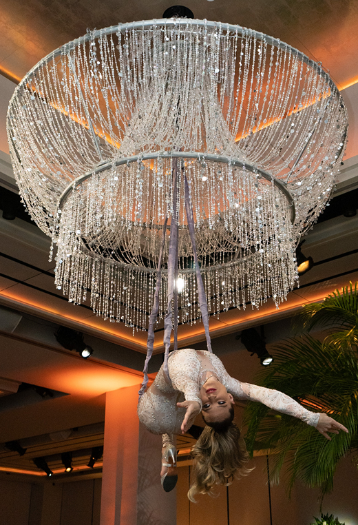 Woman in aerial silks suspended below a large crystal chandelier, arms outstretched.