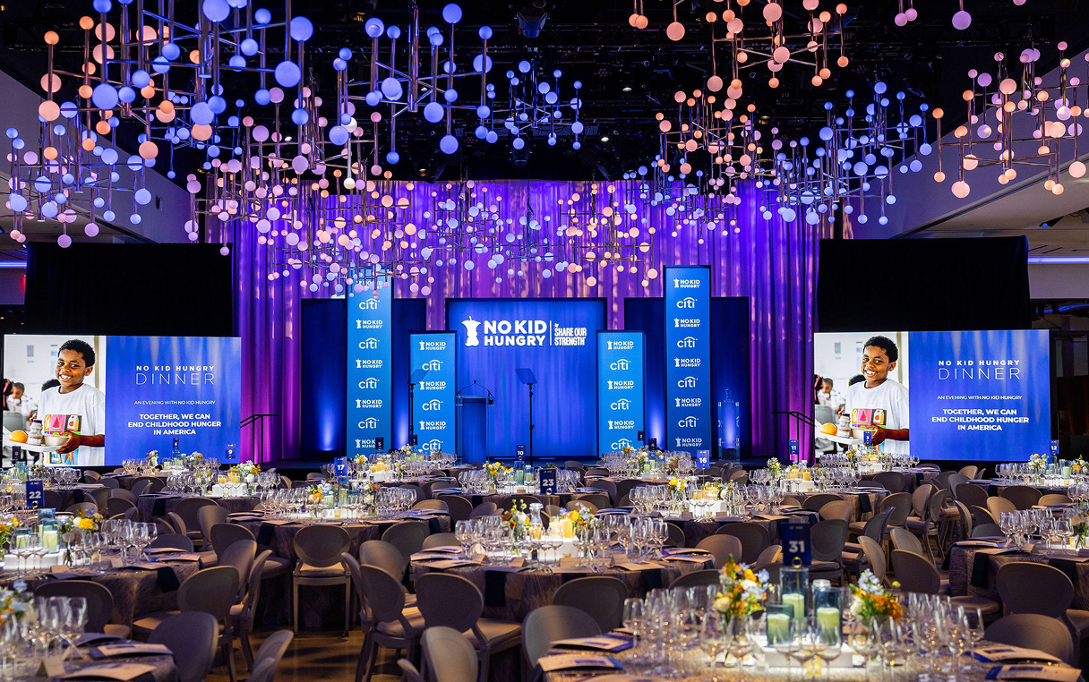Stage setup with large screens, tables with floral centerpieces, and decorative lighting.