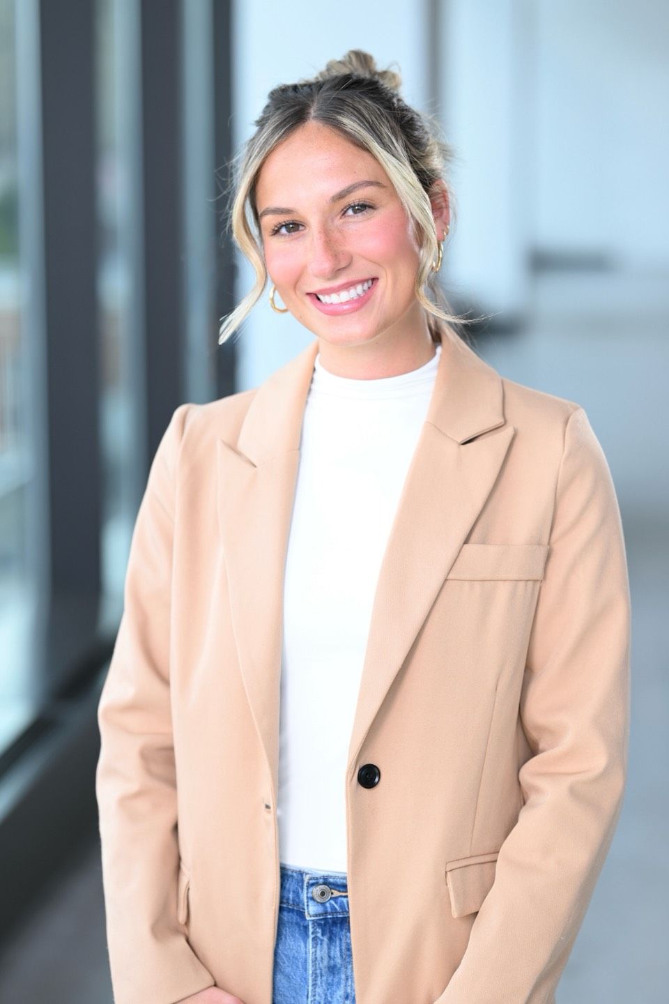 Woman with blonde hair smiling, wearing a tan blazer and white shirt, indoors near a window.