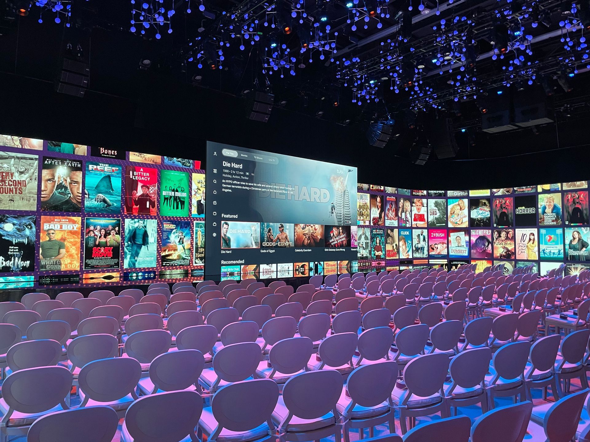Auditorium with rows of chairs facing a large screen displaying movie posters. Neon lighting highlights the space.