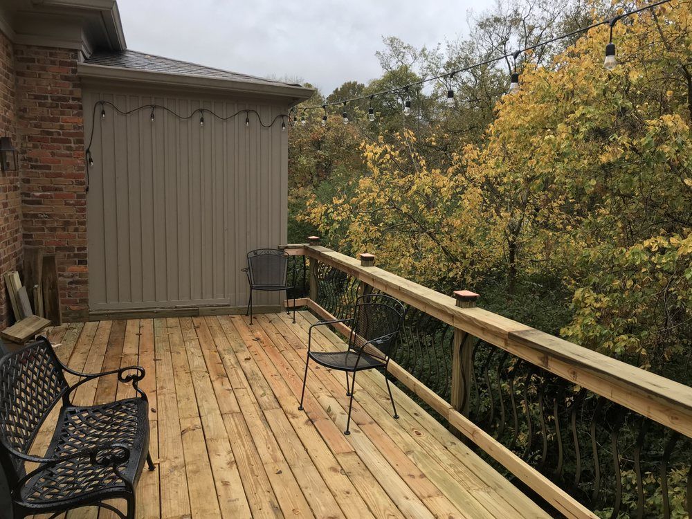 A wooden deck with a black metal bench and chair, attached to a brick house, overlooking trees with autumn leaves.