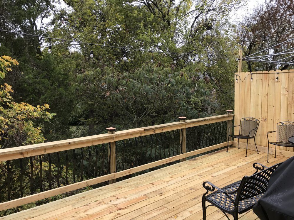 A newly built wooden deck featuring a railing with black metal balusters and a privacy wall, overlooking lush green trees.