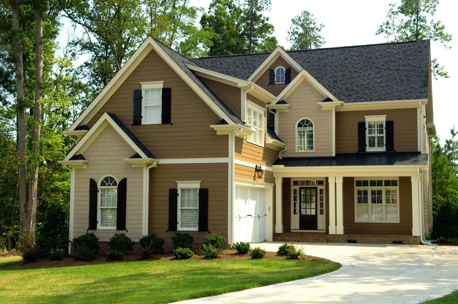 Suburban two-story tan house with white trim, black shutters, and a curved driveway surrounded by trees.