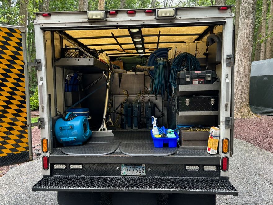 The open cargo area of a work truck, packed with organized equipment, tools, and blue supplies against a wooded backdrop.
