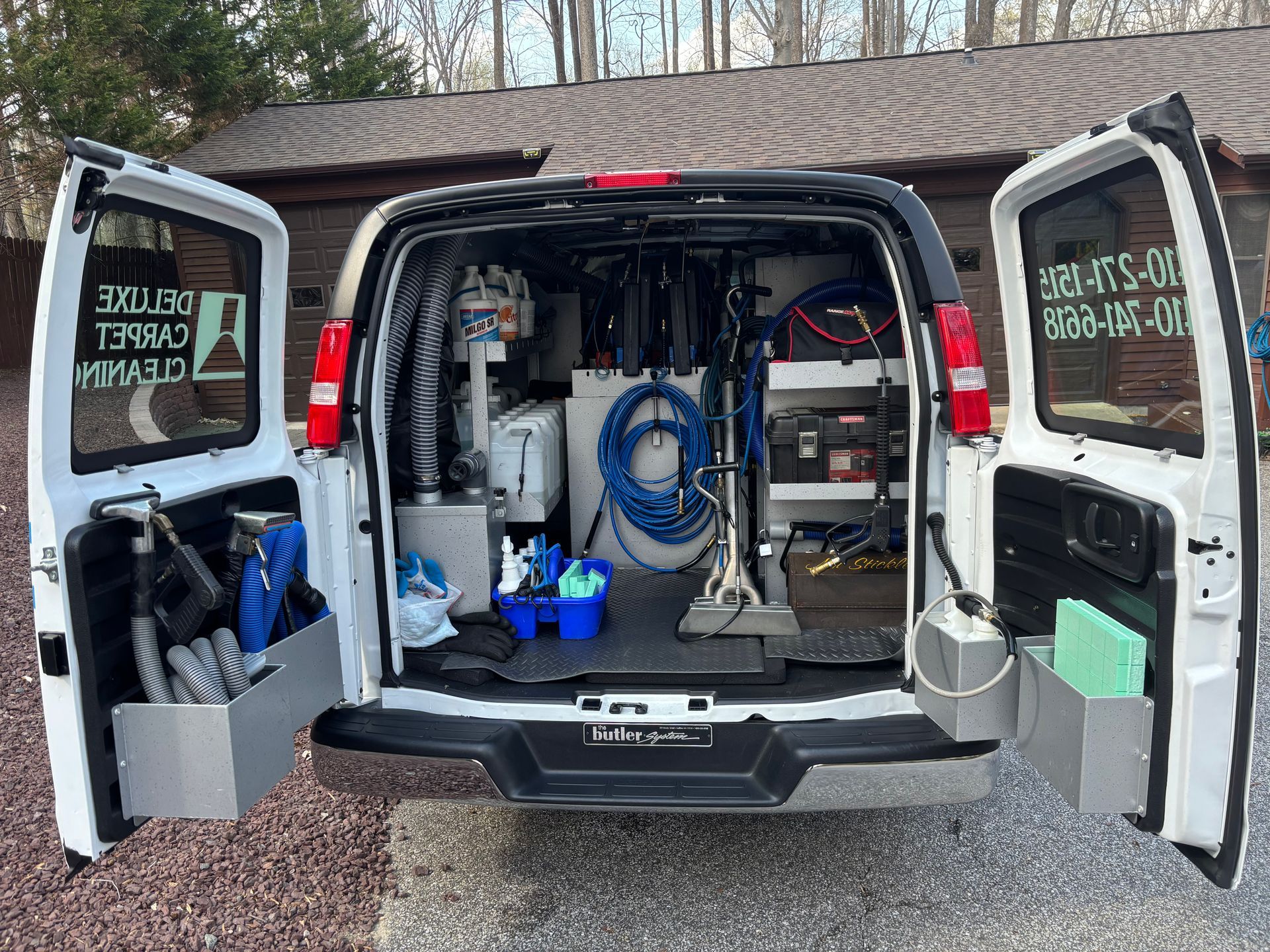 The open rear doors of a white van reveal organized carpet cleaning equipment, hoses, and containers in a driveway.