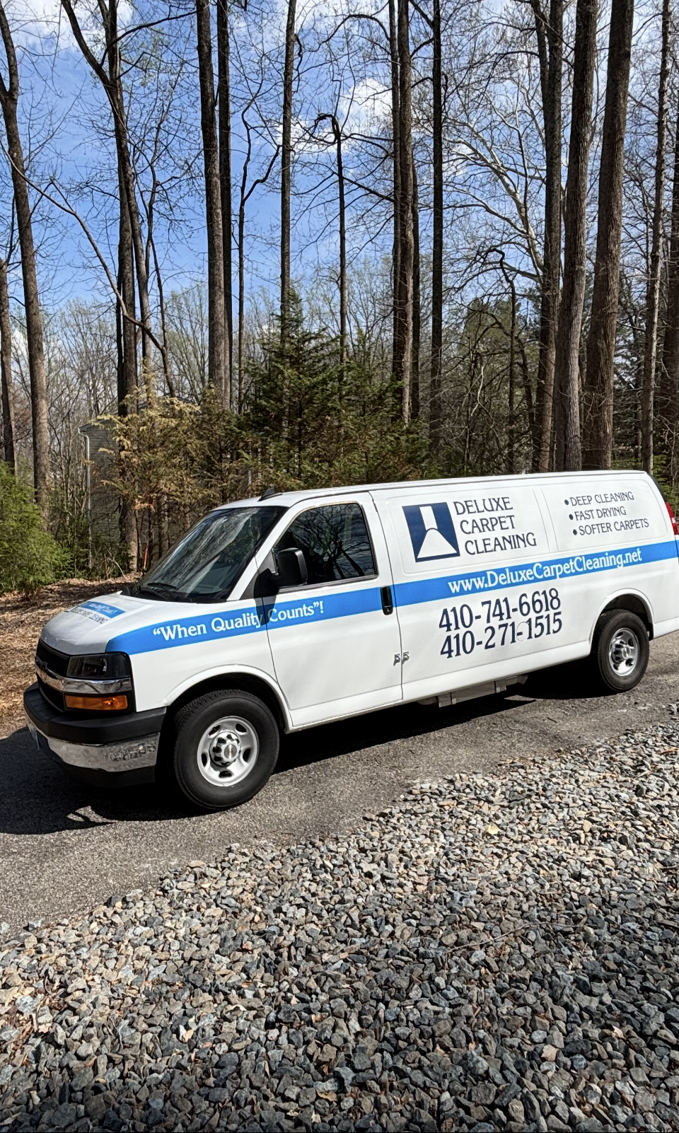 A white work van parked on a gravel path in a wooded area, displaying business branding and contact information.