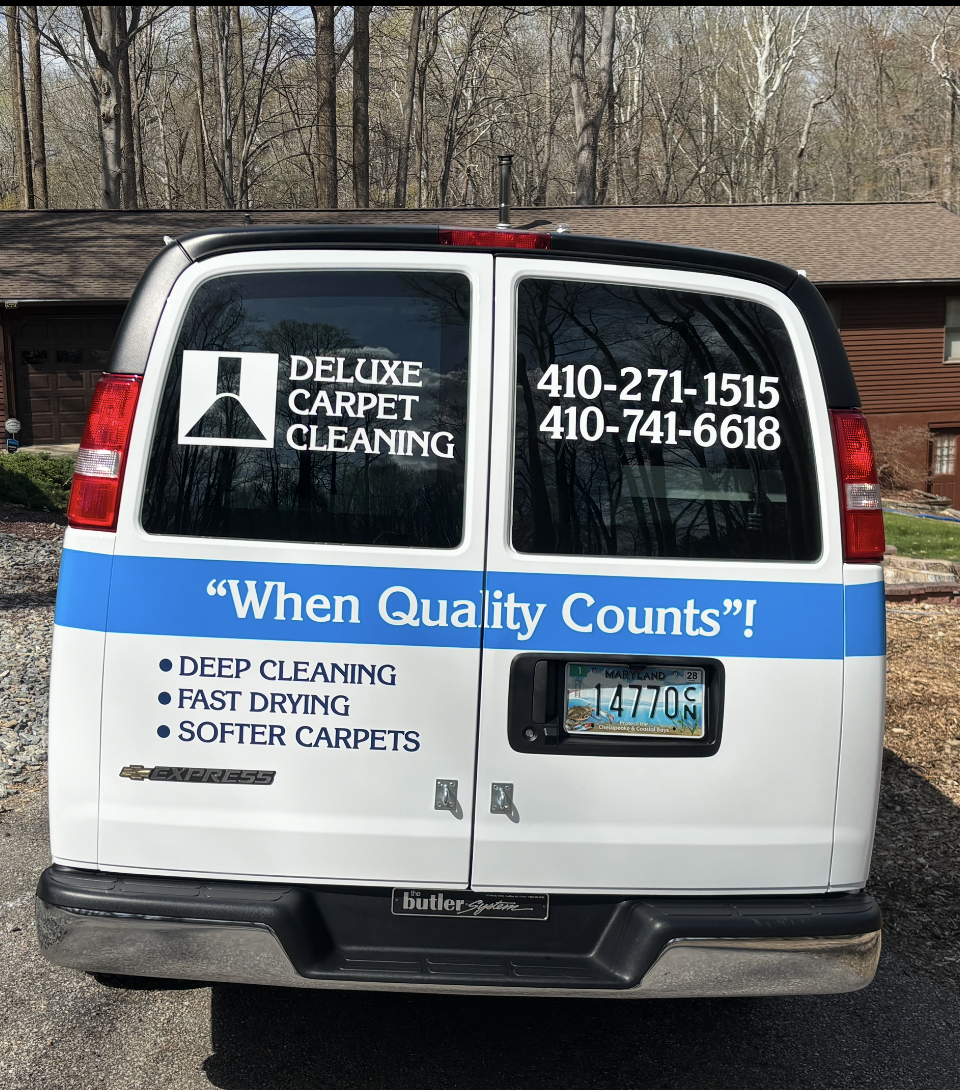 A white van with Deluxe Carpet Cleaning branding, phone numbers, and services printed on the back rear doors.