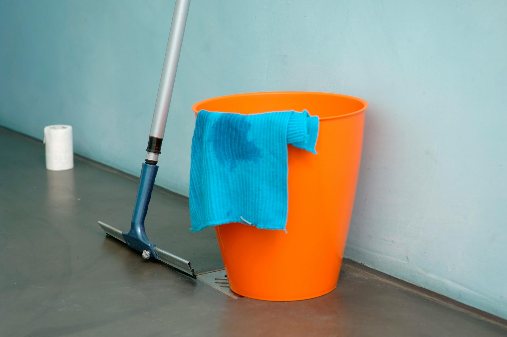 A blue squeegee, a bright orange bucket with a blue cloth draped over it, and a roll of paper towels on a grey floor.