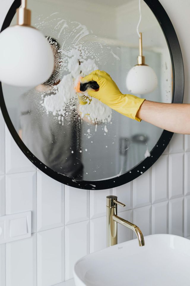 A person wearing a yellow rubber glove cleans a round mirror above a bathroom sink with white cleaning foam.