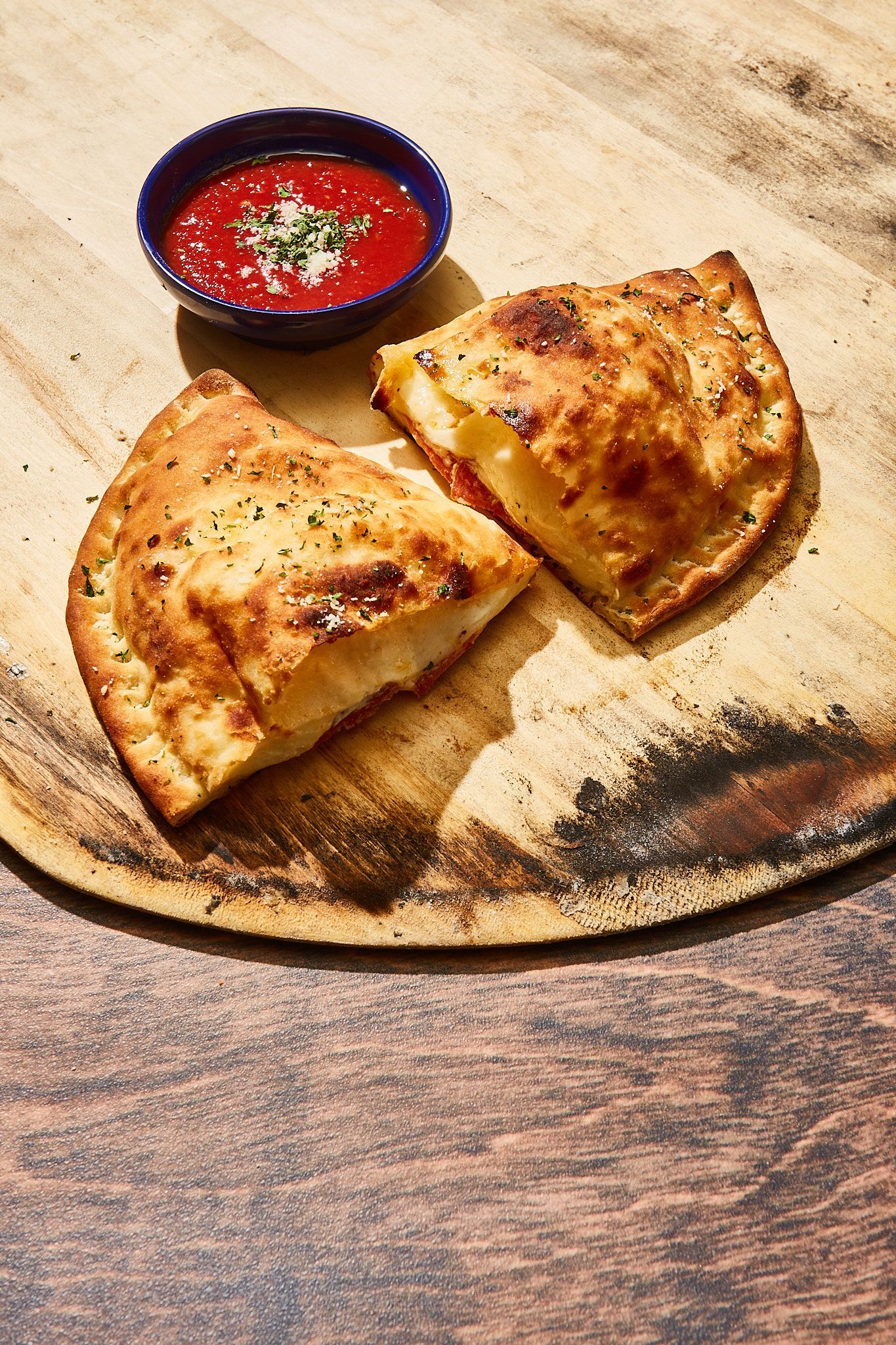 Two slices of calzone on a wooden cutting board with a bowl of tomato sauce.