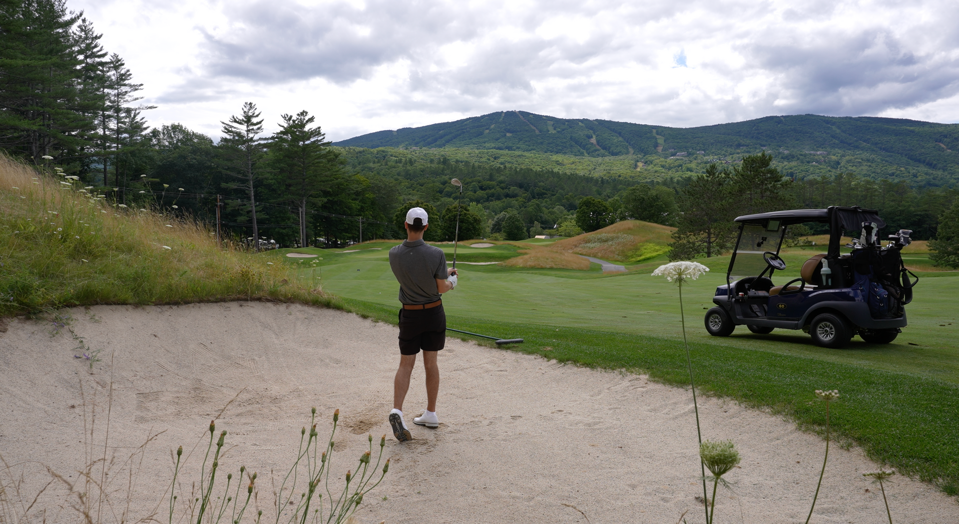 A man is standing in a bunker on a golf course next to a golf cart.