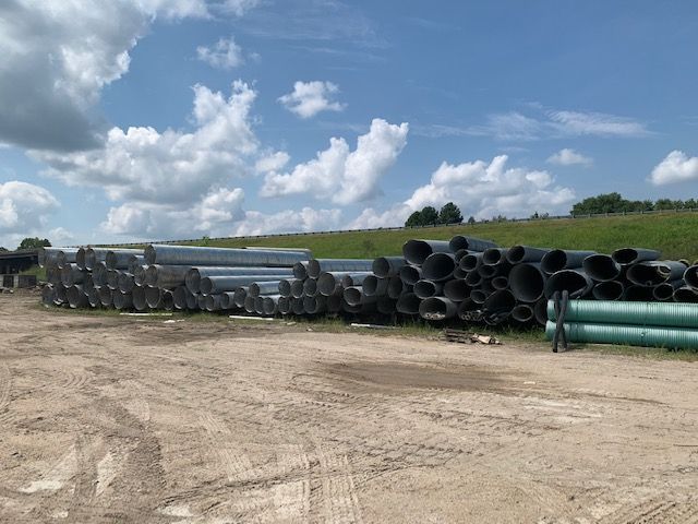Stacks of large black and green pipes on a dirt lot under a cloudy blue sky