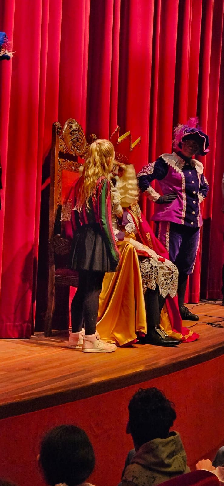 Een toneelvoorstelling met een persoon op een troon en twee mensen die ernaast staan. Rode gordijnen vormen de achtergrond.