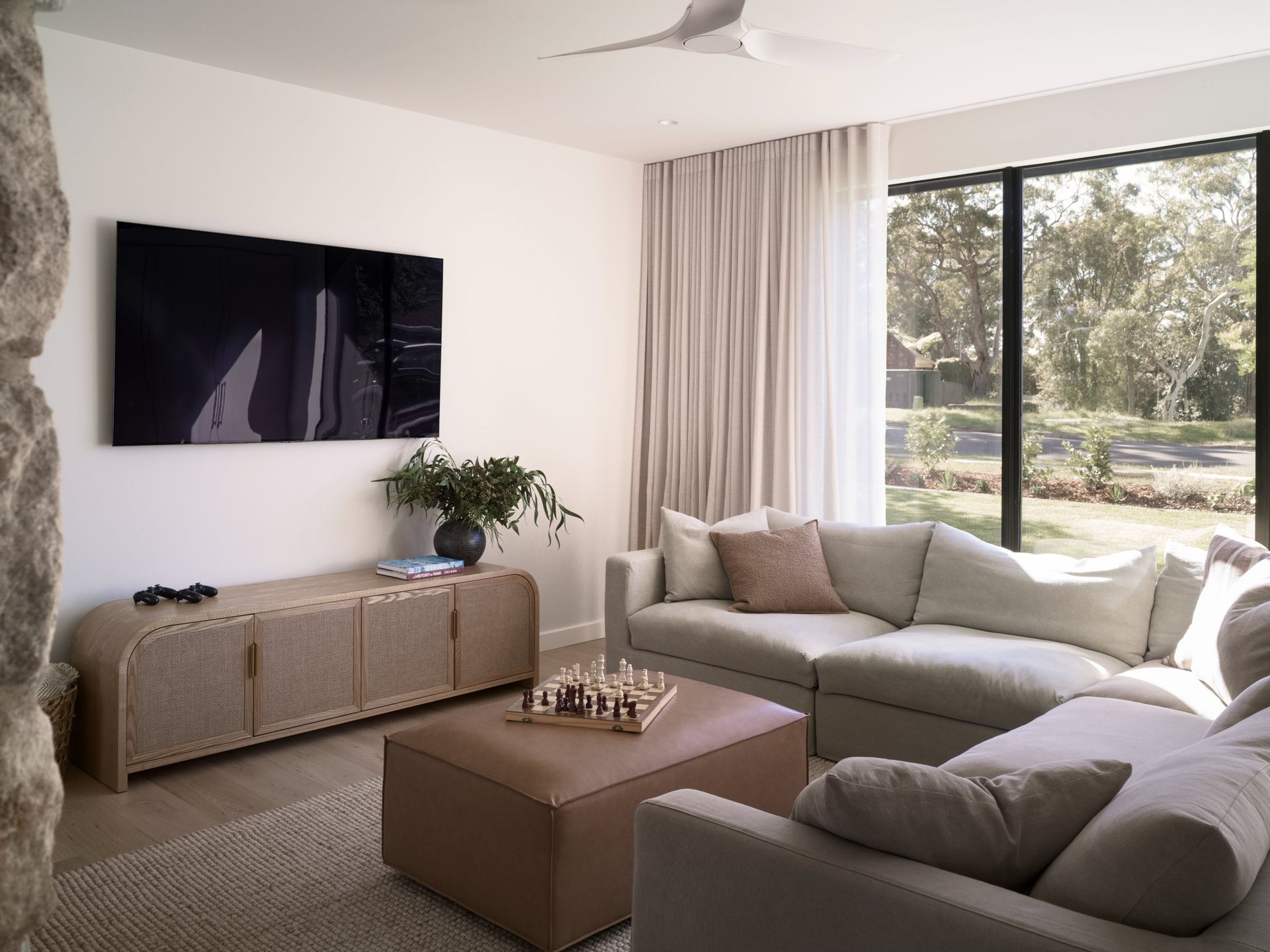 Modern living room with beige sectional sofa, wall TV, wicker console, and large window view of trees — Everyday Interiors in Helensburgh, NSW
