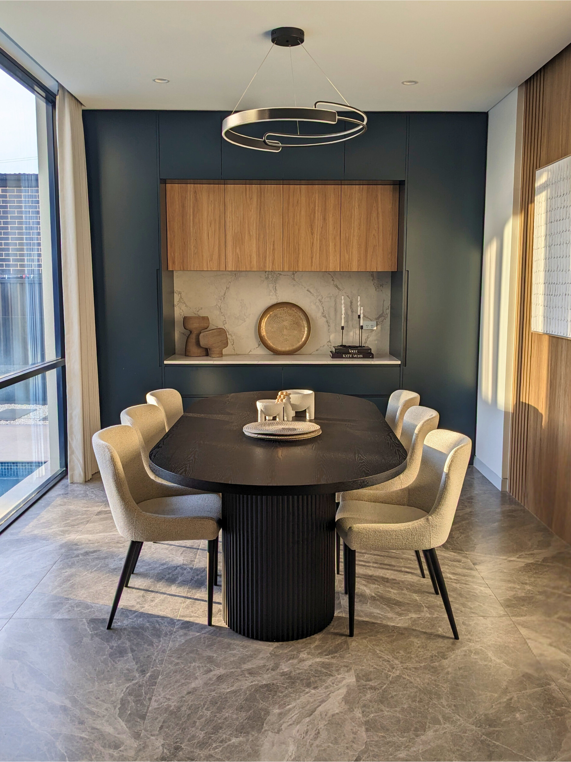 Modern dining room with a round dark table, beige chairs, blue walls, and a wood cabinet nook — Everyday Interiors in Helensburgh, NSW