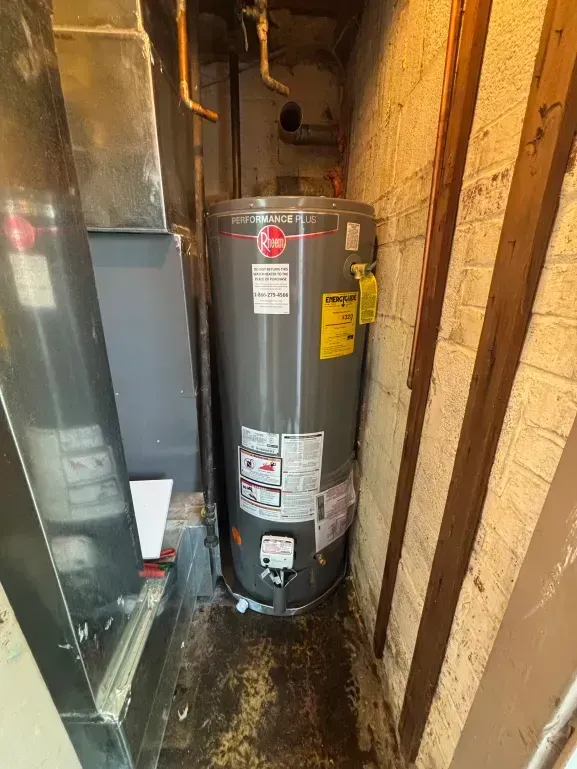 Water heater in a narrow basement space, next to a furnace and brick wall.