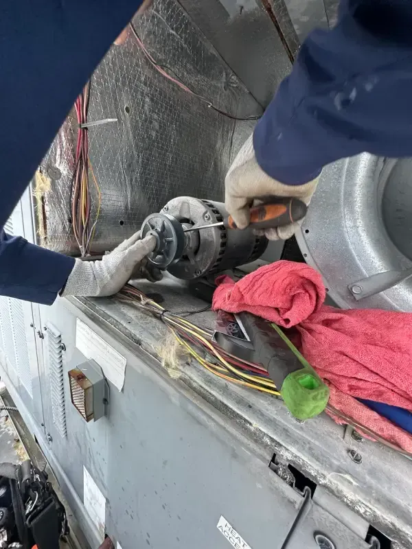 Two people in gloves work on an HVAC motor with tools, likely for repair, on a rooftop unit.