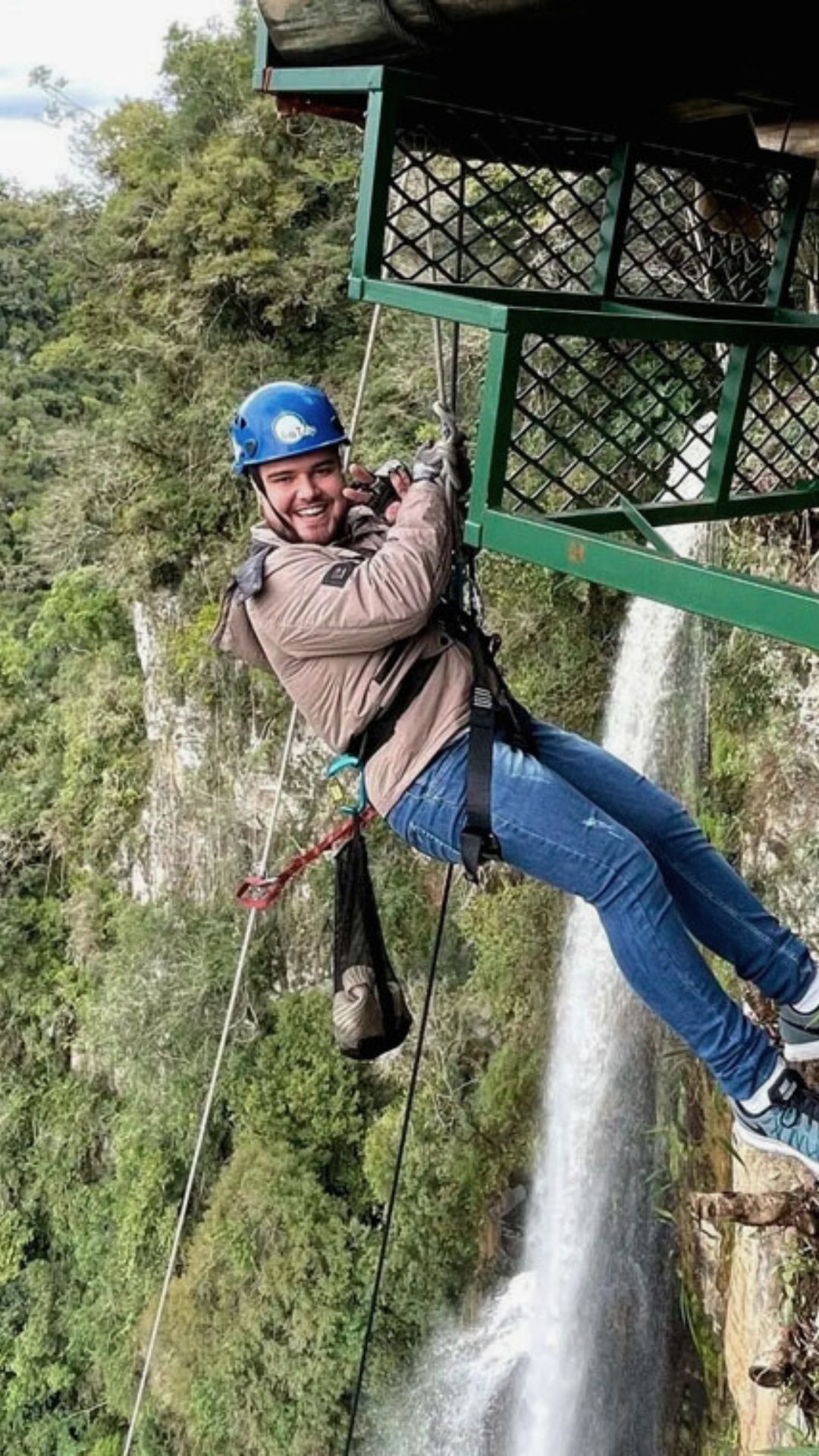 Pessoa praticando rapel perto de uma cachoeira, usando capacete, sorrindo e feliz, com folhagem verde ao redor.