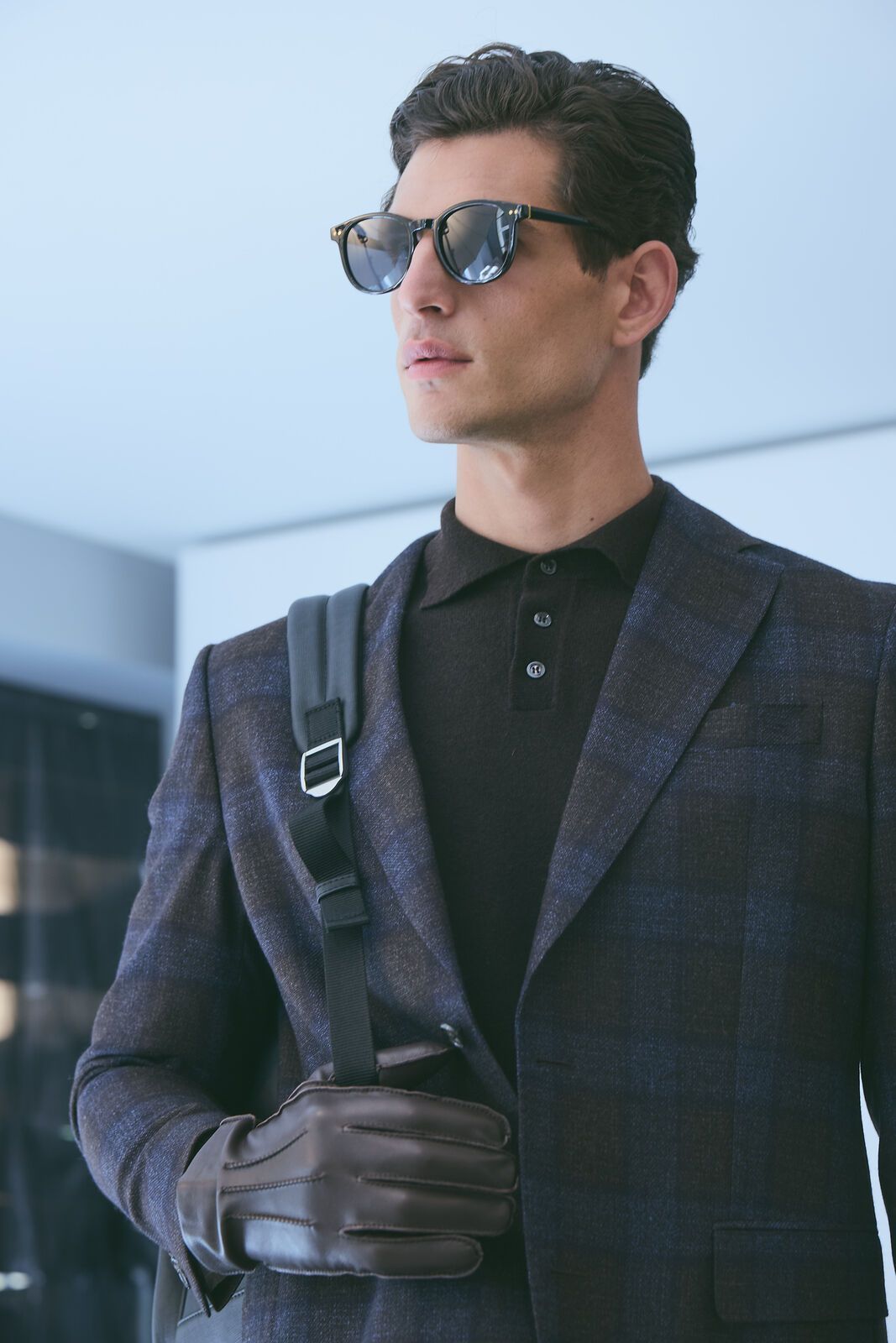Man wearing sunglasses, navy blazer, and backpack, holding gloves, standing indoors.