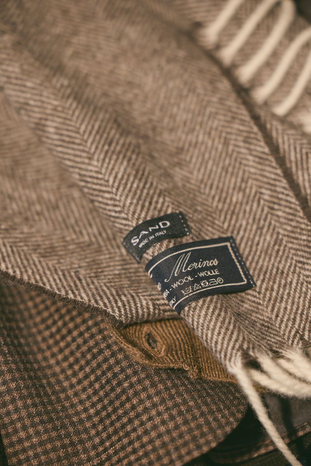Close-up of a brown herringbone-patterned scarf with a fringed edge and a fabric tag.