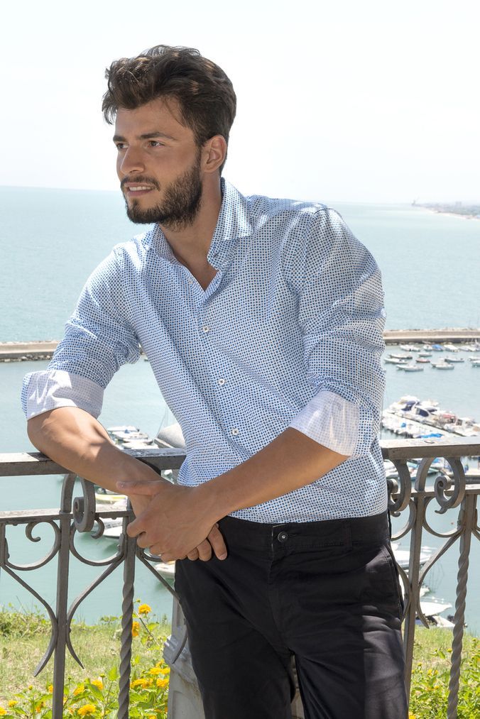 Man leaning on railing, smiling, wearing light blue patterned shirt and black pants, overlooking ocean with boats.