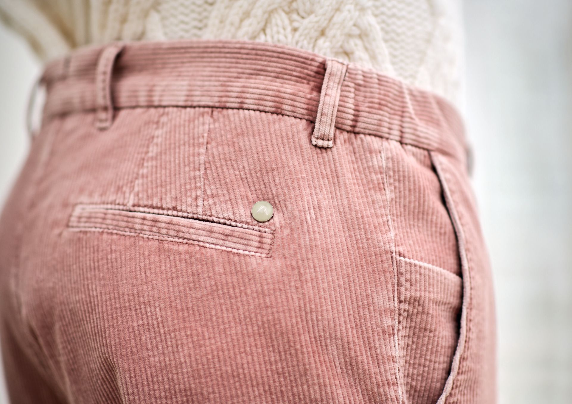 Pink corduroy pants with a back pocket and button detail. A cream-colored sweater is visible.